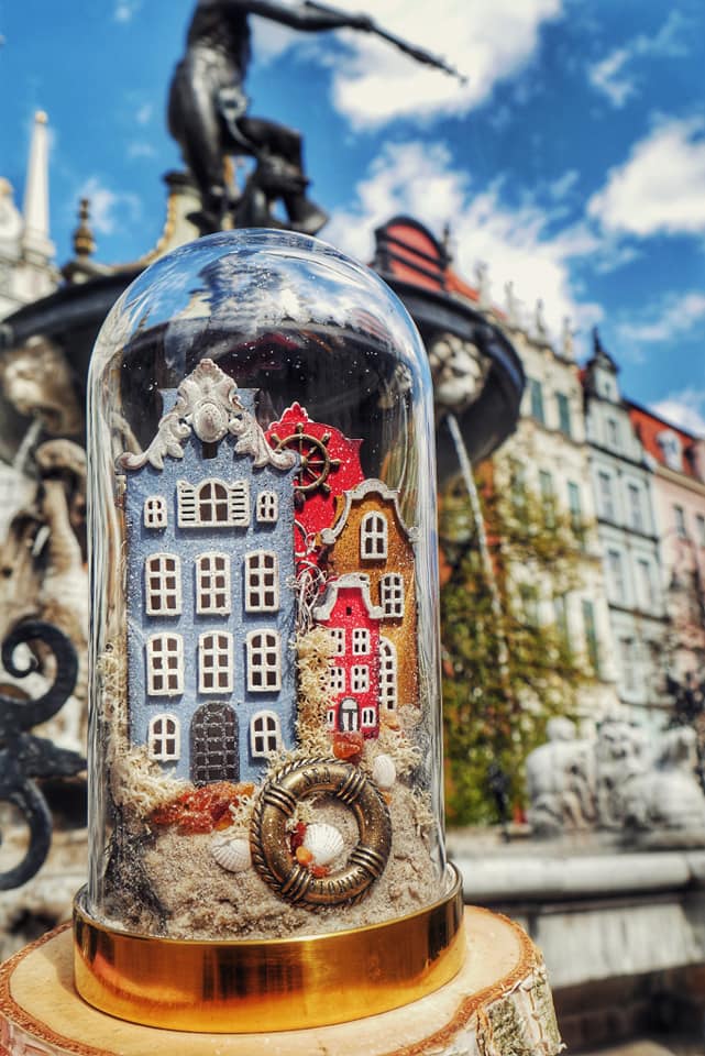 Glass dome housing a miniature colorful village with blue, red, and orange houses on a sandy base, set on a gold stand in a plaza.