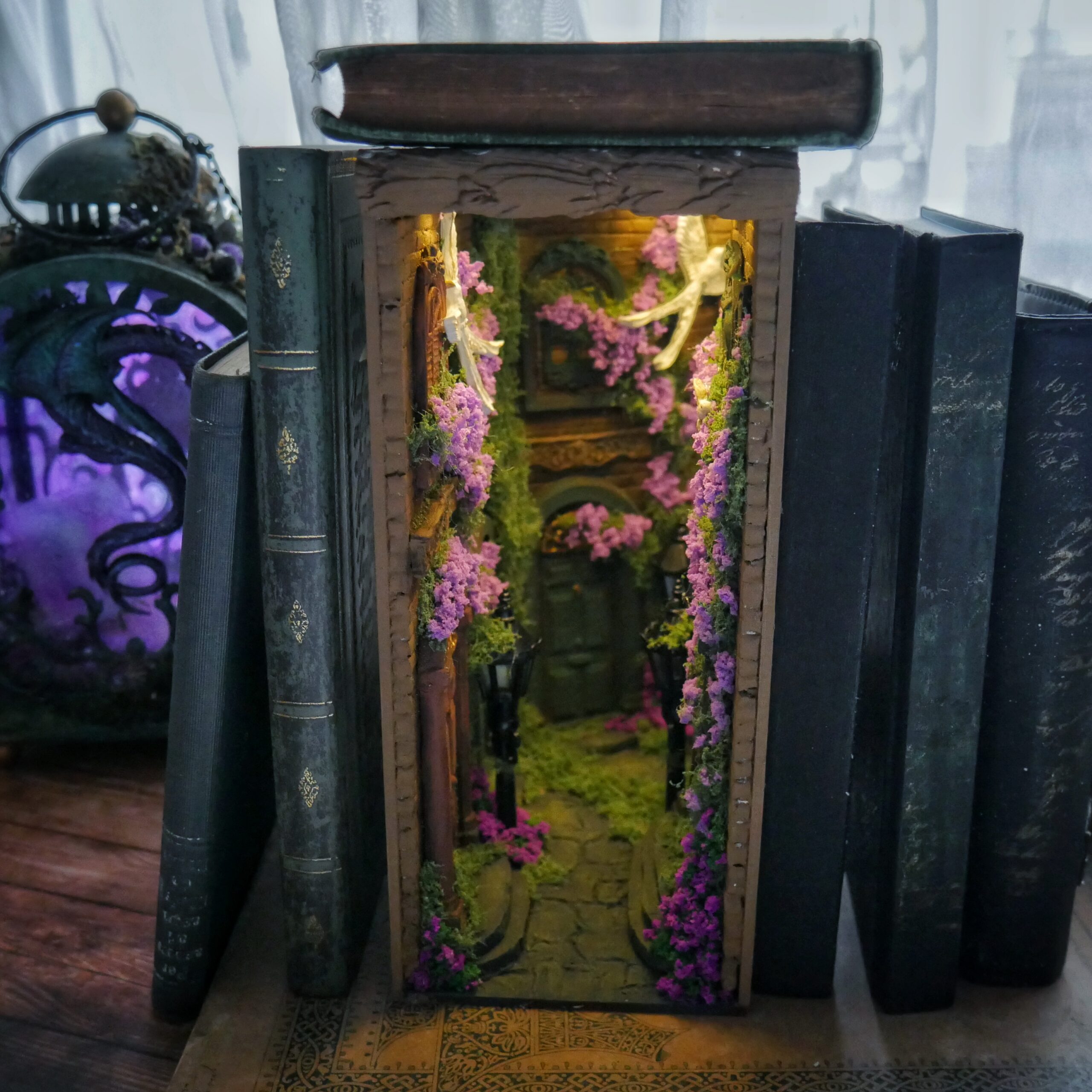 Small wooden display cabinet opened to reveal a miniature garden with purple flowers, lit from within.